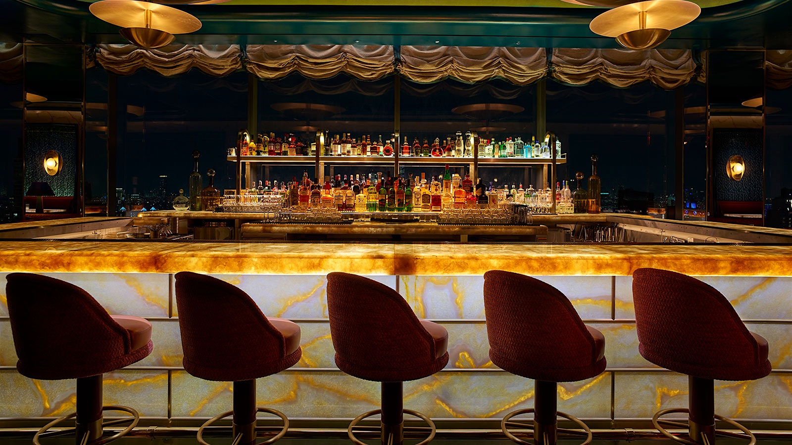  Una mirada nocturna al bar de Nubeluz con detalles dorados y esmeraldas y lujosos taburetes de bar color burdeos